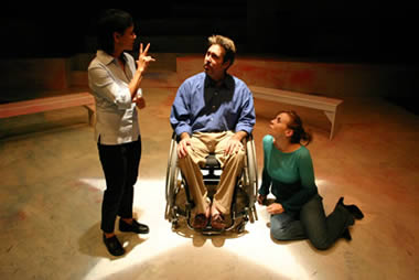 A man in a wheelchair between a woman on the floor leaning on him and another wo